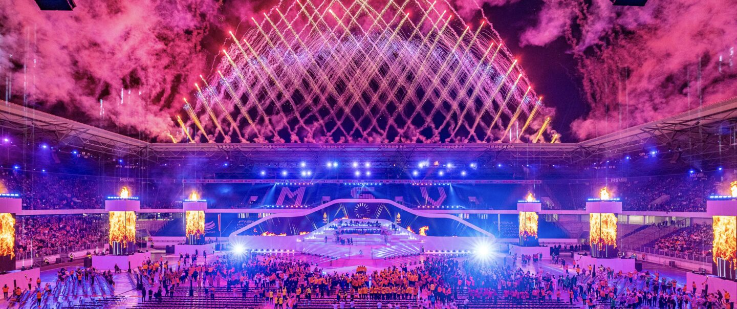Großes Stadionpublikum mit Blickrichtung Himmel, erleuchtet von Feuerwerksreflexionen.