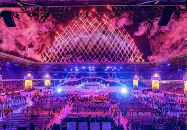 Großes Stadionpublikum mit Blickrichtung Himmel, erleuchtet von Feuerwerksreflexionen.