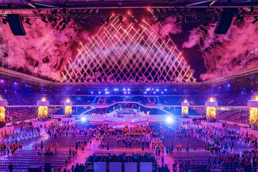 Großes Stadionpublikum mit Blickrichtung Himmel, erleuchtet von Feuerwerksreflexionen.