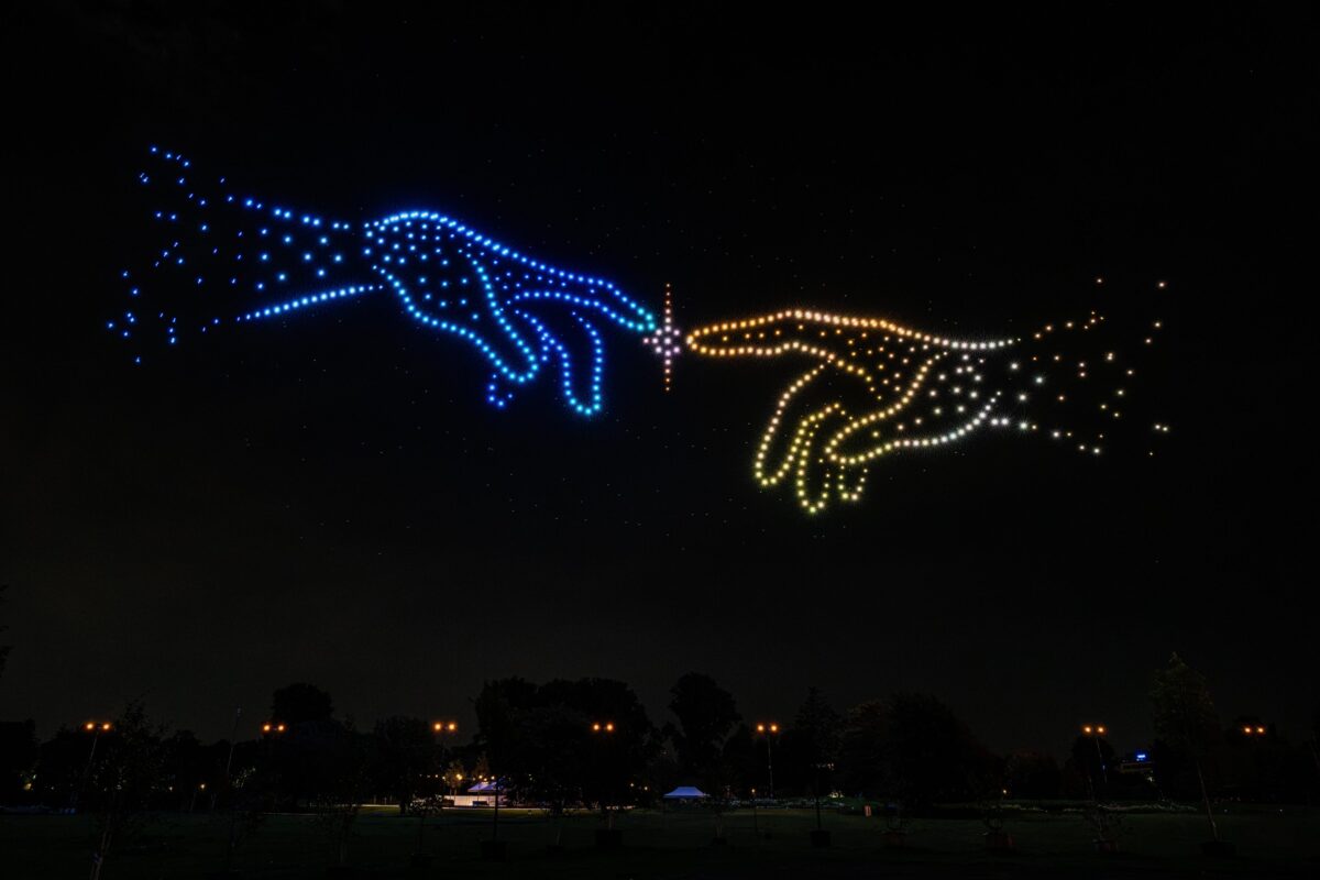 Zwei Hände aus Lichtdrohnen nähern sich bei der Landesgartenschau Neuss 2026 an, während zwischen den Fingerspitzen ein leuchtender Funke entsteht